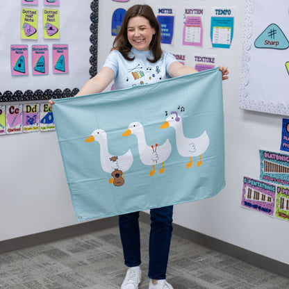 A woman in a classroom setting holds up a light blue fabric banner featuring three cartoon ducks playing musical instruments.