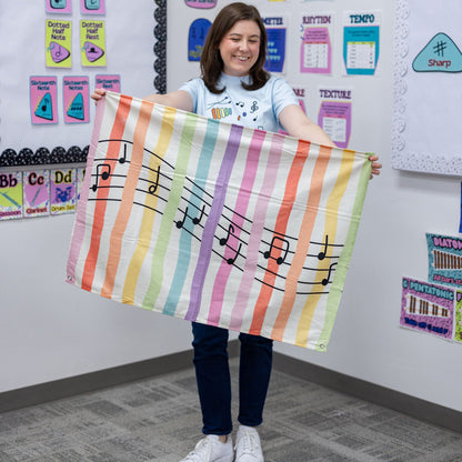 A woman stands in a classroom holding up a large striped fabric banner with black musical notes. The banner has vertical stripes in rainbow colors including pink, orange, yellow, green, blue, and purple, with black musical notes printed across the center.