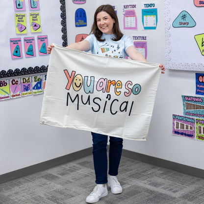 A woman stands in a classroom holding up a white banner that reads 'You are so Musical' in colorful lettering. The room has colorful educational posters and charts on the walls.