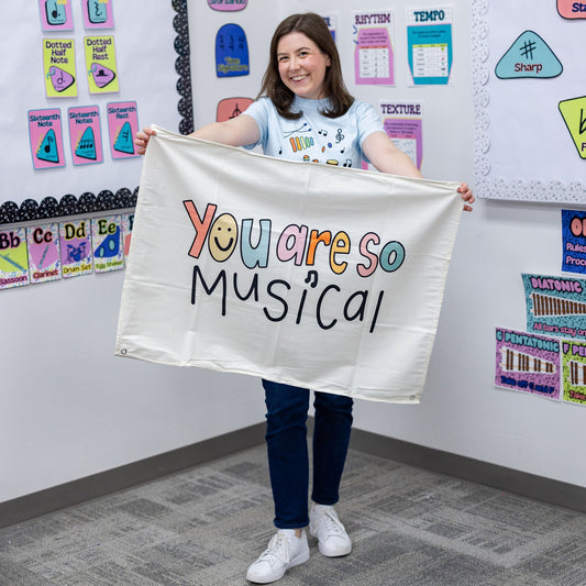 A woman stands in a classroom holding up a white banner that reads 'You are so Musical' in colorful lettering. The room has colorful educational posters and charts on the walls.