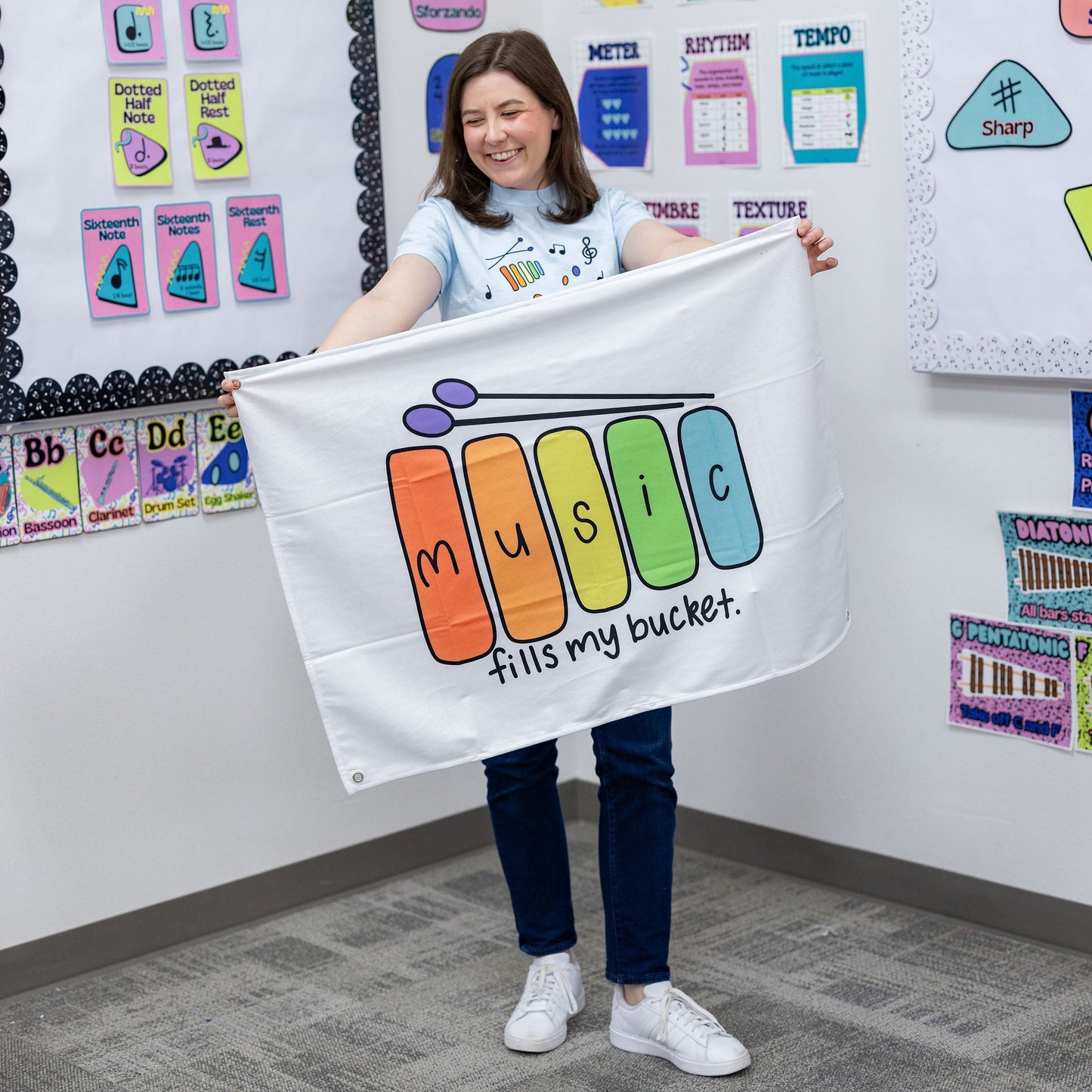 A woman in a classroom setting holds up a white banner that reads 'music fills my bucket.' with colorful letters spelling 'music' in a piano-key style.