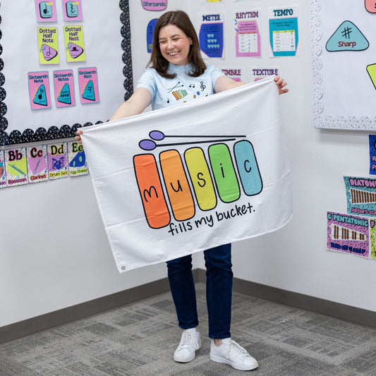 A woman in a classroom setting holds up a white banner that reads 'music fills my bucket.' with colorful letters spelling 'music' in a piano-key style.