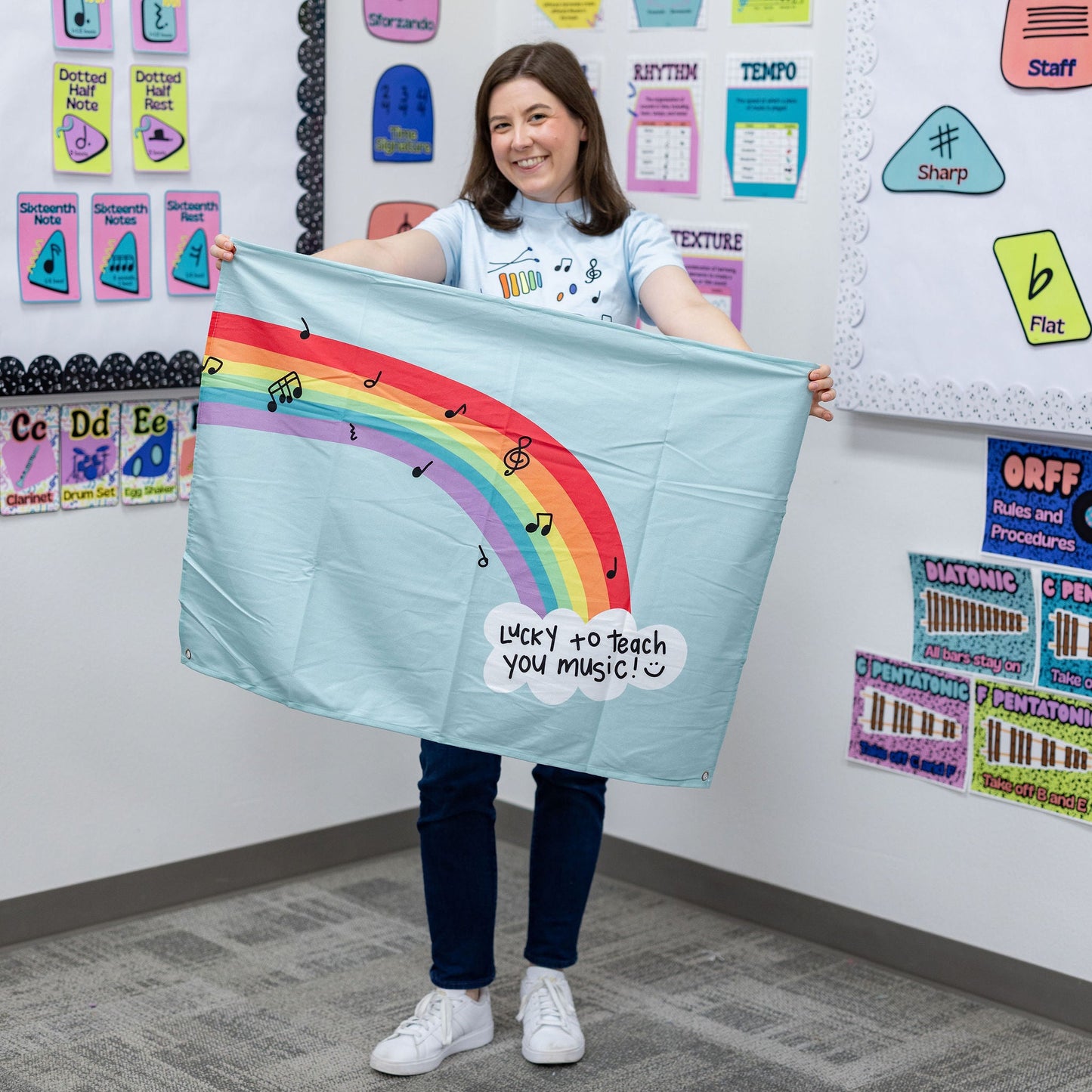 A woman stands in a classroom holding up a light blue fabric banner with a colorful rainbow graphic and the text 'Want to teach you magic!'. The background shows educational posters and charts on a white wall.