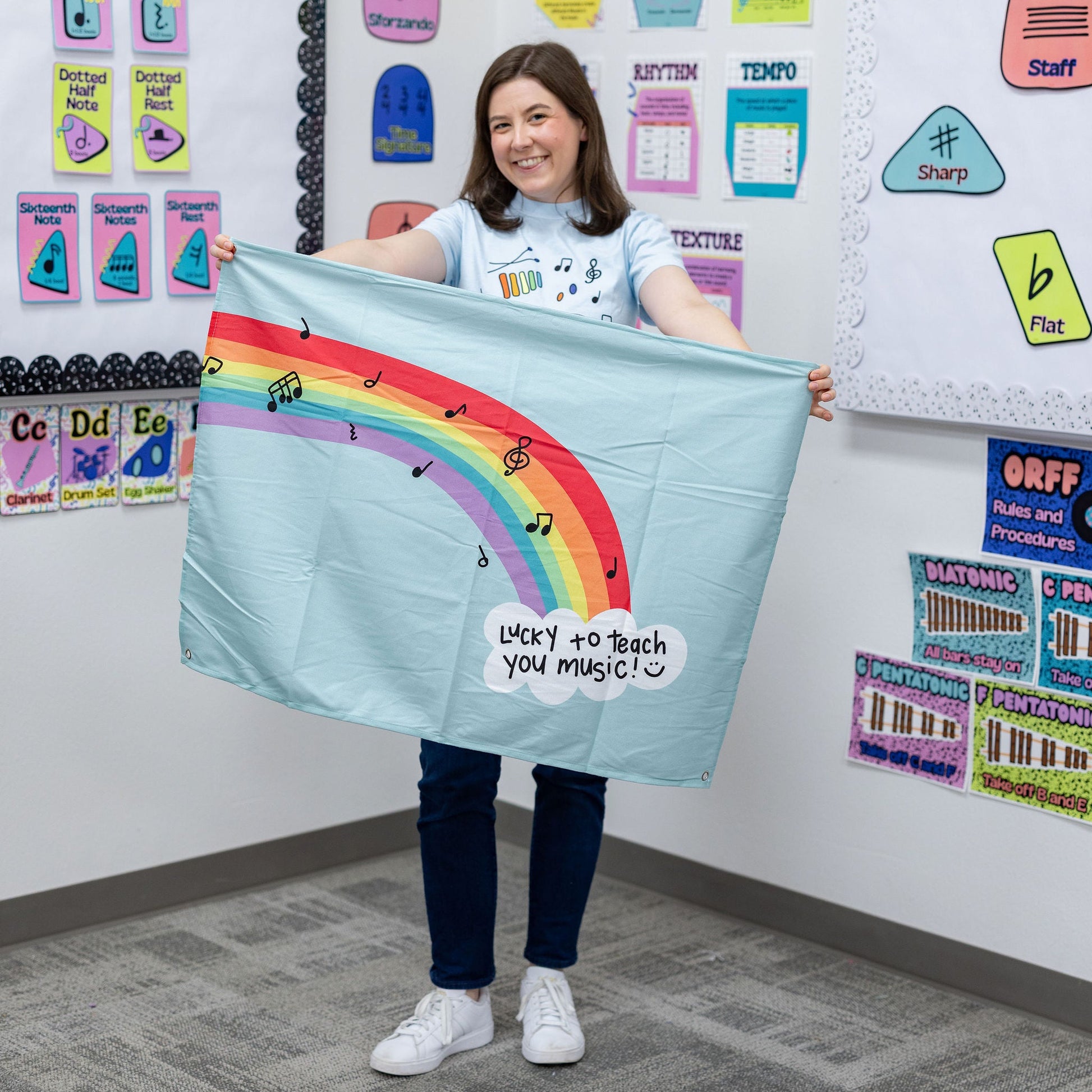 A woman stands in a classroom holding up a light blue fabric banner with a colorful rainbow graphic and the text 'Want to teach you magic!'. The background shows educational posters and charts on a white wall.