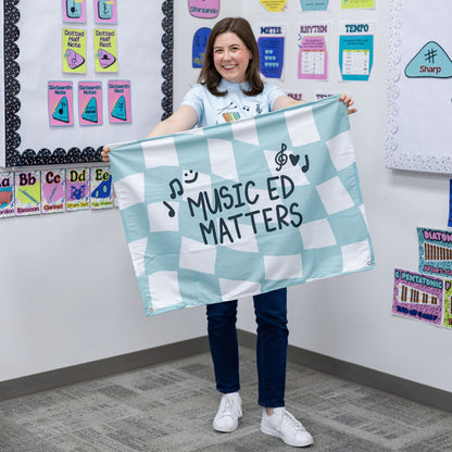 A smiling woman stands in a classroom holding a light blue and white checkered banner that reads 'MUSIC ED MATTERS' with music note graphics. The room has colorful educational posters and charts on the walls.