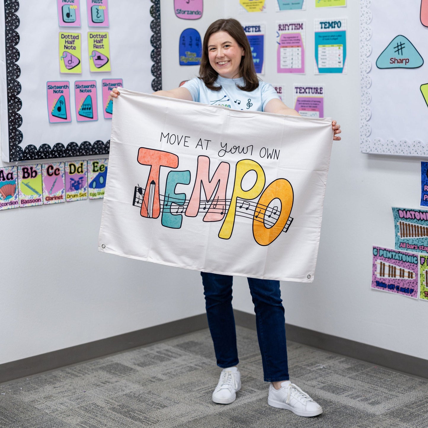 A woman stands in a classroom holding up a white banner that reads 'MOVE AT YOUR OWN TEMPO' in colorful lettering. The room has educational posters and charts on the walls.
