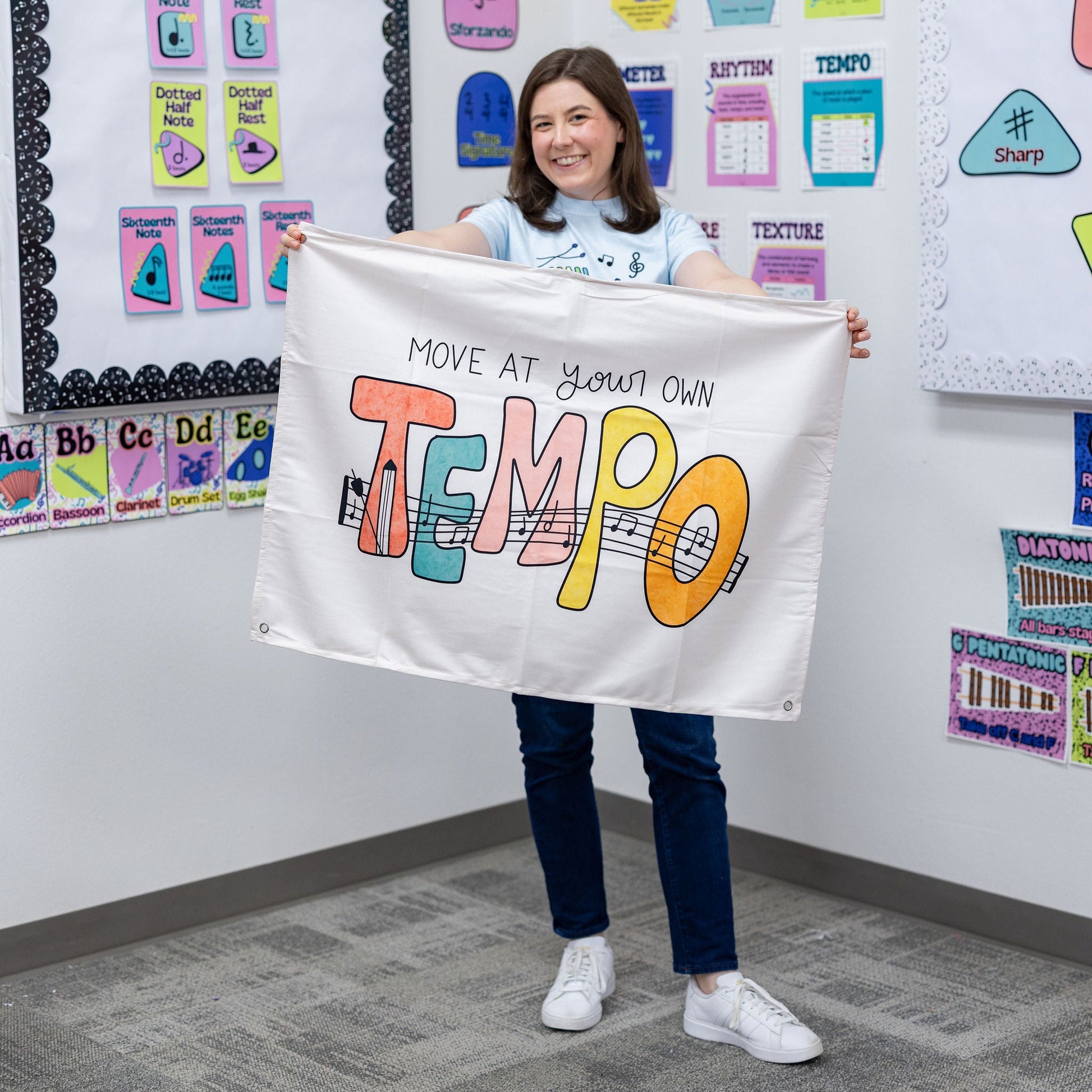 A woman stands in a classroom holding up a white banner that reads 'MOVE AT YOUR OWN TEMPO' in colorful lettering. The room has educational posters and charts on the walls.