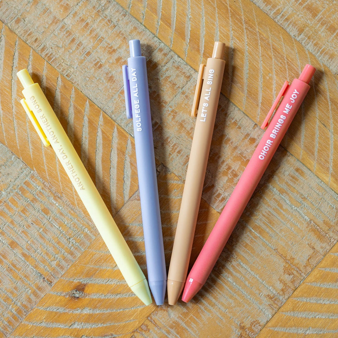 Four pastel-colored pens in yellow, blue, beige, and pink arranged in a fan shape on a wooden surface.