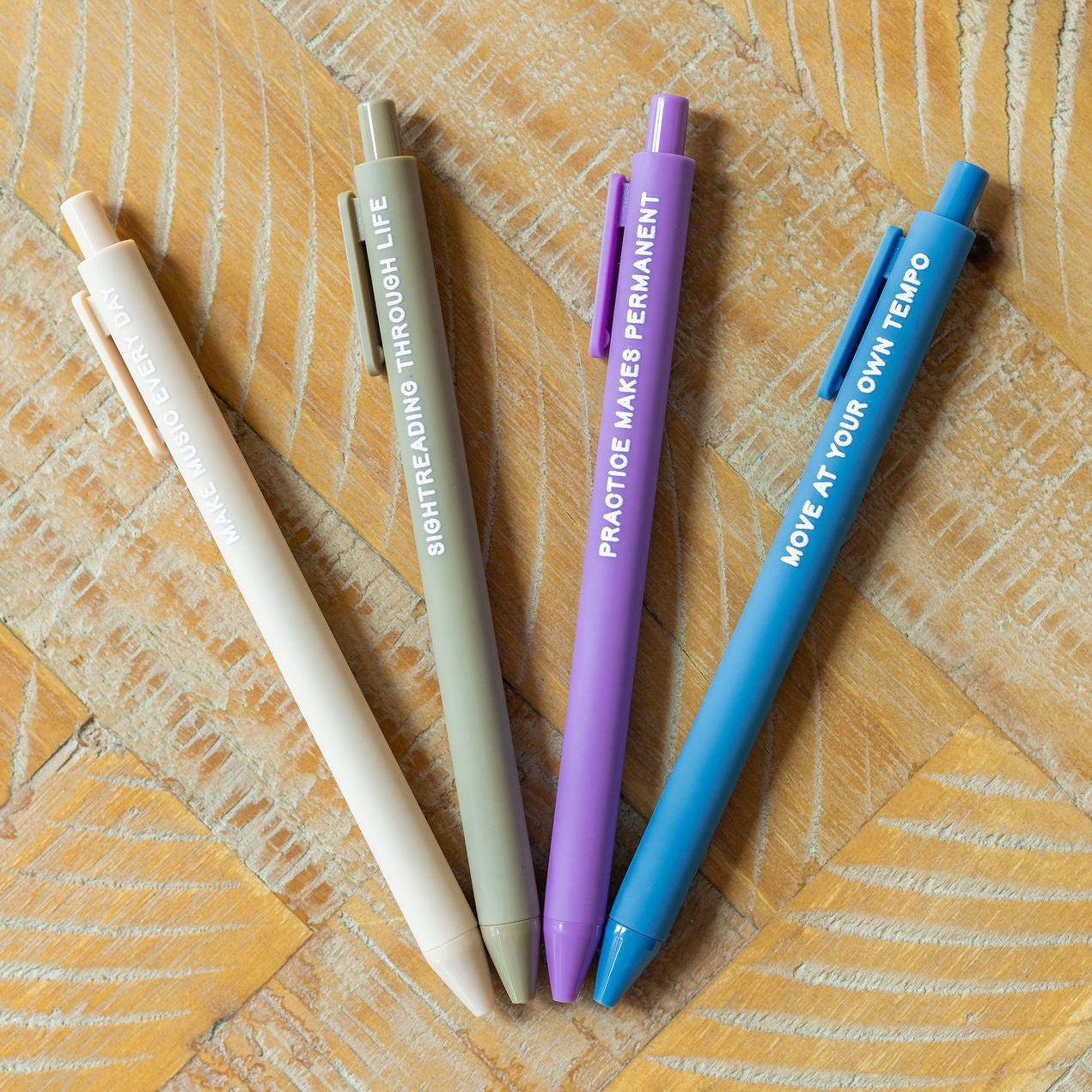Four retractable gel pens in beige, gray, purple, and blue, arranged in a fan shape on a light wood background. Each pen features white text with a motivational quote.