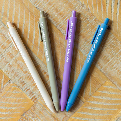Four retractable gel pens in beige, gray, purple, and blue, arranged in a fan shape on a light wood background. Each pen features white text with a motivational quote.