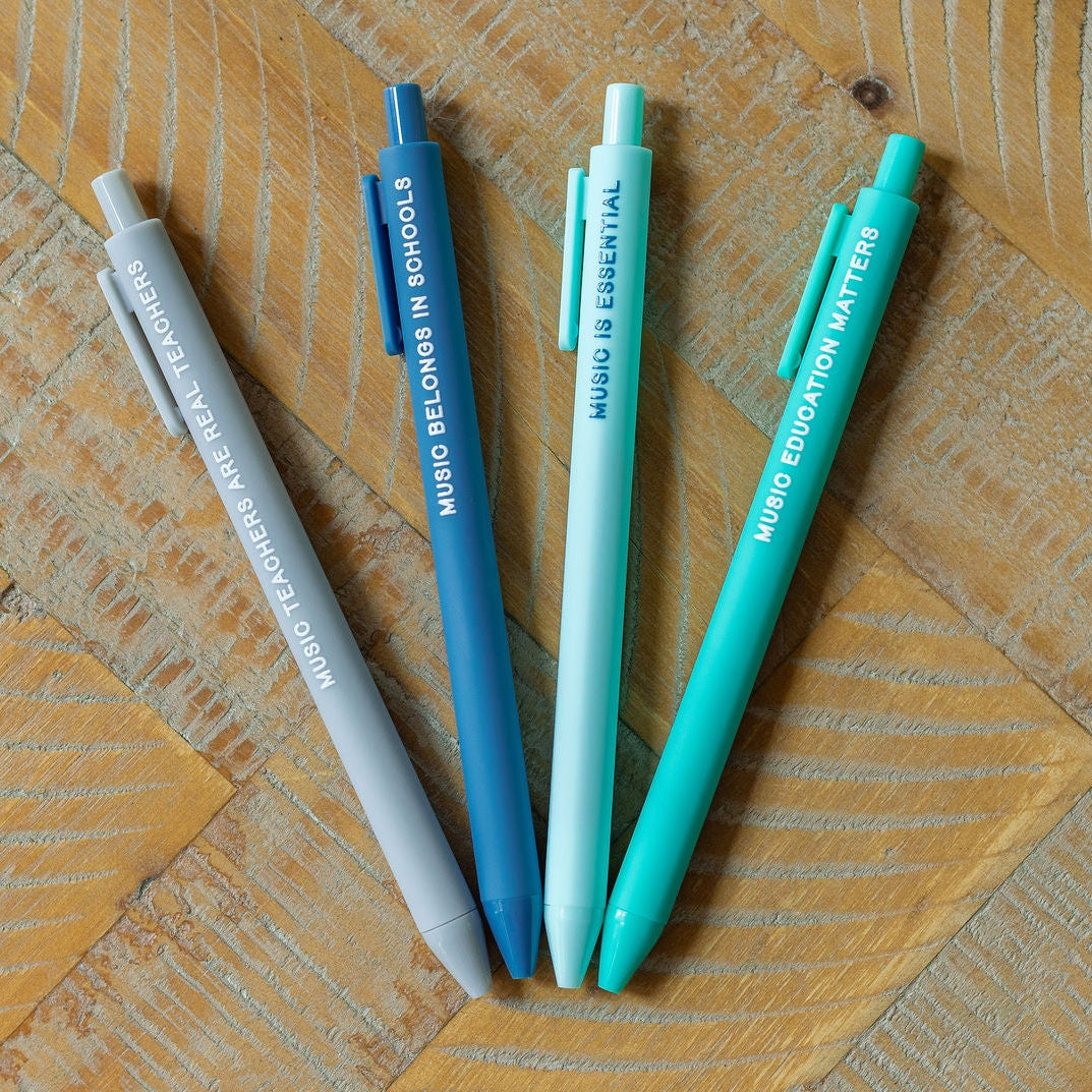 Four retractable ballpoint pens in light gray, blue, white, and mint green, arranged on a wooden surface with a visible grain pattern.