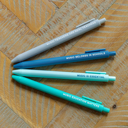 Four Micron pens in gray, blue, white, and teal, printed with 'MUSIC BELONGS IN SCHOOL' and 'MUSIC IS ESSENTIAL' / 'MICRON EDUCATION PEN', arranged diagonally on a wooden surface.