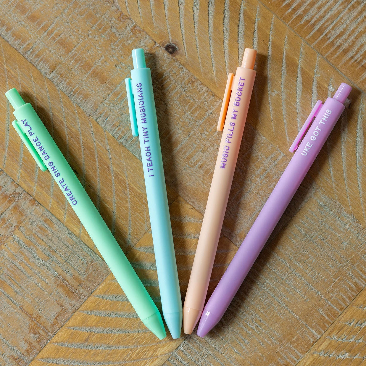 Four pastel-colored retractable pens arranged in a fan shape on a wooden surface. The pens are mint green, light blue, peach, and lavender, each printed with a school-themed phrase.