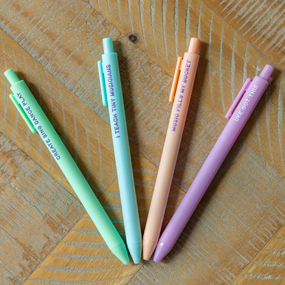 Four pastel-colored retractable pens arranged in a fan shape on a wooden surface. The pens are mint green, light blue, peach, and lavender, each printed with a school-themed phrase.